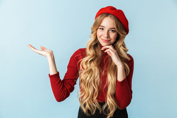 Image of caucasian blond woman 20s wearing red beret smiling and holding copyspace on palm while standing, isolated over blue background in studio