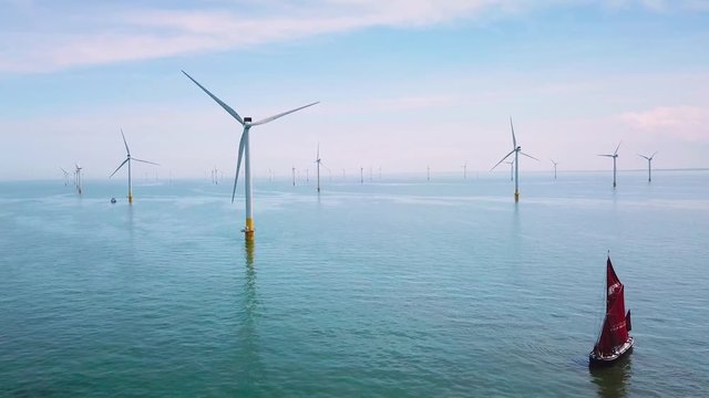 A flat bottomed sailing barge sailboat moves up the Thames River Estuary in England amidst numerous wind turbine windmills.
