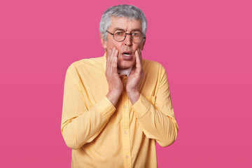 Headshot of stupefied terrified thin man wears yellow shirt, keeps hands on cheeks. Surprised elderly male with glasses against rose wall. Person knows bad news. People and emotions concept.