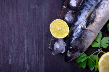 Fresh mackerel on a black chopping board surrounded by ice, lime, lemon, parsley and spices