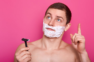 Thoughtful Caucasian man with shaving gel, raises fore finger, idea in mind, holds sharp razor in...