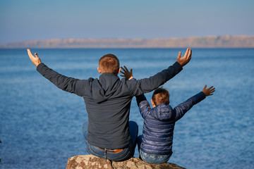 Dad and son are sitting on the bank of the lake and enjoying the atmosphere around their arms are open wide.
