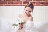 Portrait of gorgeous bride in a white dressing gown in the apartments. She is holding wedding bouquet in her hands and smiling.