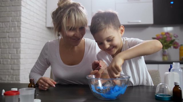 Cute preadolescent son making slime, thoroughly mixing pasty mass to desired texture with spoon. Joyful mother watching fascinating process, communicating with smiling child and having fun too