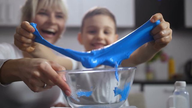 Close up handmade slime made by preadolescent son with help of loving mother in domestic kitchen. Little boy's hands stretching ready slime on camera. Blurry on background proud family. Dolly
