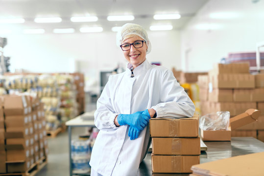 Smiling Caucasian blonde female employee in sterile uniform and with eyeglasses leaning on boxes and standing in food factory.