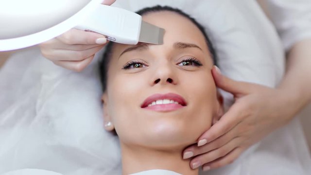 Happy Caucasian young woman enjoying facial ultrasonic peeling procedure at beauty salon