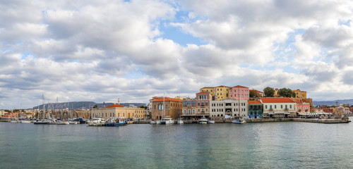 A panorama of the seaport town Chania, the island of Crete, Greece. A harbor with wooden pantons, moored yachts, ships, boats. Colorful architecture of modern and old houses. Mountains on the horizon.
