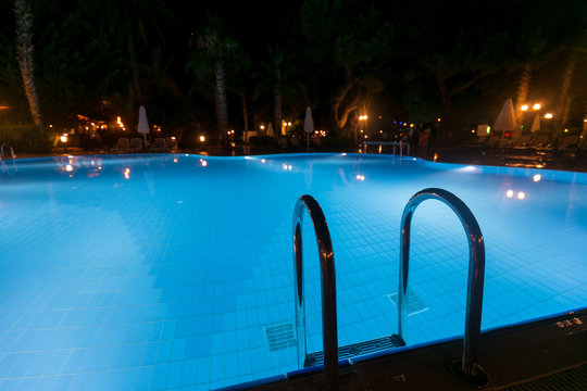 Illuminated swimming pool at night time. Grab bars ladder in the blue swimming pool at tropical resort.