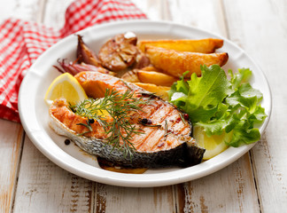 Grilled salmon steak, a portion of grilled salmon with fresh lettuce and potato wedges on a white ceramic plate on a wooden rustic table, top view, close-up.