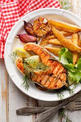 Grilled salmon steak, a portion of grilled salmon with fresh lettuce and potato wedges on a white ceramic plate on a wooden rustic table, top view, close-up.