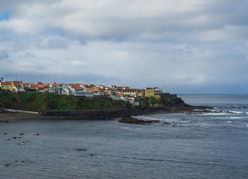 Colorful small coastal village houses on rocky lava shore above Atlantic ocean with sea water waves, cloouds and moody sky, Maia village, Sao Miguel, Azores islands, Portugal