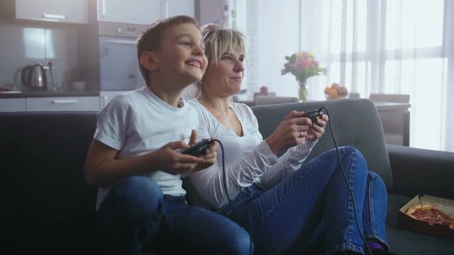 Adorable little boy competing with pretty adult mother in video game sitting on couch in domestic interior. Cheerful family having fun together playing home console using joysticks