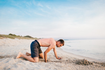 Young topless barefoot man in shorts digging hole by hands on beach beyond sea in summer evening....