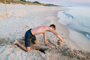 Young topless barefoot man in shorts digging hole by hands on beach beyond sea in summer evening....