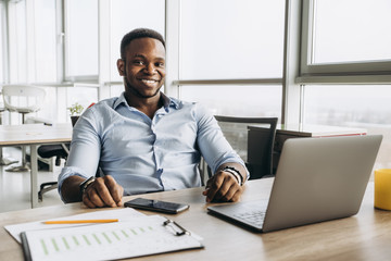 Fototapeta na wymiar Cheerful successful modern man of African-American appearance, holding a high position in his company, working in a laptop at his workplace