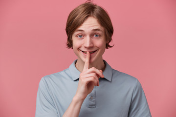 Fototapeta premium Happy smiling young guy keeps fore finger on lips, calls to keep a secret, do not tell anyone, keep quiet, do not make noise, demonstrates silence gesture, isolated over pink background