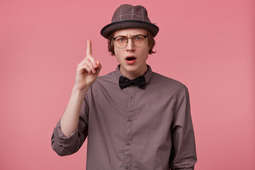 Serious young smartly dressed guy holding hand up pointing index finger upwards, looking camera through glasses moralizing, comment on issues of right and wrong, makes moral lecture, pink background