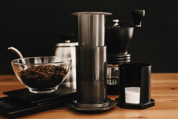 Aeropress, steel kettle, scales, manual grinder, coffee beans on wooden table and black background. Alternative coffee brewing method set. Stylish accessories and items for alternative coffee
