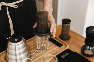 Professional barista pouring hot water in aeropress, alternative coffee brewing method. Hands holding steel kettle and aeropress, glass cup, scales, coffee beans on wooden table