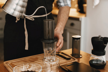 Hands holding steel kettle and aeropress, glass cup, scales, grinder on wooden table. Professional barista pouring hot water in aeropress, alternative coffee brewing method.