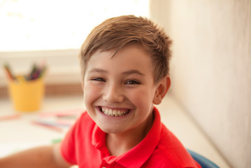 cute happy brunette child 9-10 years old in red shirt drawing at home at table with colored pencils