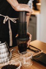 Professional barista preparing coffee by aeropress alternative method, brewing process. Hands on aeropress and glass cup, scales, manual grinder, coffee beans, kettle on wooden table