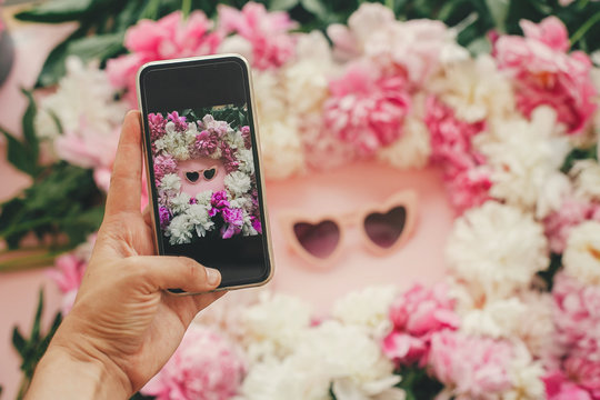 Hand holding phone and taking photo of pink heartshaped sunglassess in stylish pink and white peonies frame on pink paper flat lay. Instagram blogging. Summer vacation