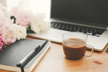 Freelance concept. International womens day. Stylish working place. Stylish laptop, coffee cup, notebook, pen,  pink and white peonies on wooden table with space for text.