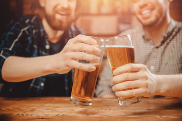 Hand with glasses of beer foam, guys ring mugs, say toast, chin-chin, drink to health. Young bearded men in background blurred.