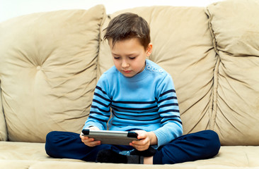 Serious kid sits on a sofa with his legs and plays on the electronic gadget at home. Little boy in blue sweater sitting on a soft couch and playing on a tablet