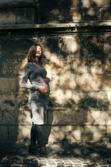 Stylish pregnant woman holding hands on baby bump and relaxing in sunny light in city street. Happy young parent, mom hugging belly, and enjoying moment. Motherhood concept. True happiness