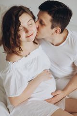 Happy pregnant couple relaxing on white bed and holding belly bump. Happy young husband kissing his smiling wife and hugging baby belly.  Stylish pregnant family in white at home. True happiness