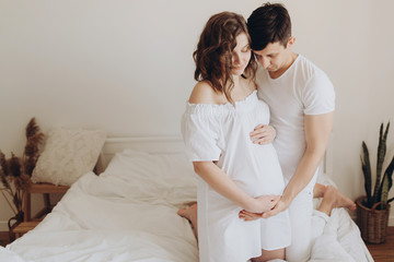 Happy young pregnant couple holding belly bump on white bed. Stylish pregnant family, mom and dad in white, relaxing at home and hugging belly. Fertility concept. Moment of true happiness