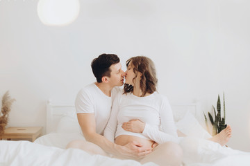Obraz na płótnie Canvas Happy young husband kissing his smiling pregnant wife and hugging belly bump on white bed. Stylish pregnant couple in white relaxing and holding belly at home. Fertility concept. True happiness
