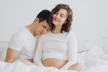 Happy young pregnant couple holding belly bump on white bed. Stylish pregnant family, mom and dad in white, relaxing at home and hugging belly. Fertility concept. Moment of true happiness