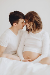 Happy young husband kissing his smiling pregnant wife and hugging belly bump on white bed. Stylish pregnant couple in white relaxing and holding belly at home. Fertility concept. True happiness