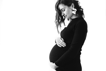 Fashionable pregnant mom, hugging belly. Stylish Motherhood concept. Space for text. Happy young pregnant woman in stylish black dress and modern earring, holding belly bump and posing in light