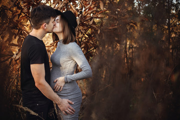 Happy young parents, mom and dad, hugging baby bump, kissing, enjoying beautiful moment at sunset. Parenthood concept. Stylish pregnant couple holding hands on belly in sunny light in autumn park.