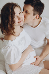 Fototapeta premium Happy young husband kissing his smiling pregnant wife and hugging belly bump on white bed. Stylish pregnant couple in white relaxing and holding belly at home. Fertility concept. True happiness