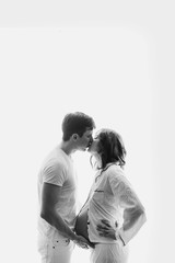 Happy young husband kissing his pregnant wife and holding belly bump. Stylish pregnant family, mom and dad in white pajamas, relaxing at home in light. Fertility concept. True happiness