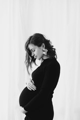 Fototapeta premium Happy young pregnant woman in stylish black dress holding belly bump and posing in light. Stylish fashionable mom, hugging belly, waiting for baby. Motherhood and fertility concept