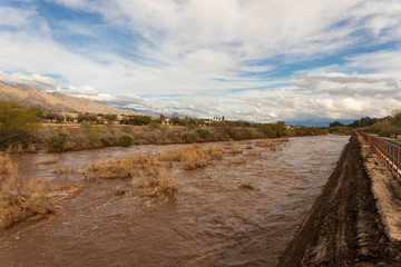 Brown water rages down normally dry riverbed under beautiful partly cloudy sky