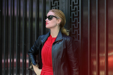 Sexy Beauty Blond hair Girl with red Lips wearing red long dress and leaf jacket is posing outdoor at urban area of the city. Luxury Woman in stylish glasses and black leaf jacket.
