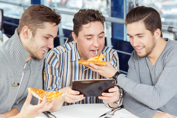 That is what friends are for. Young man giving his friend a piece of pizza while watching something on the tablet together