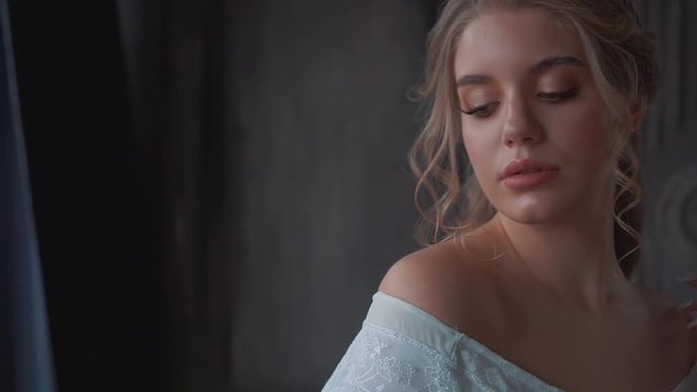 sweet lady in medieval room with blond hair and delicate skin in long gorgeous chic white dress posing for camera, cute curls of blond hair in young girl, video portrait, maid with tender nude makeup