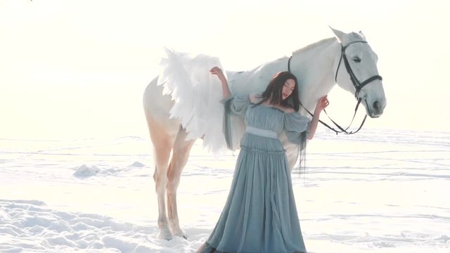 cute pretty girl hides behind white horse, touches back of warm body of pegasus and feels energy, kind fairy with dark hair in long gray vintage light dress, lady protected by magical mythical animal