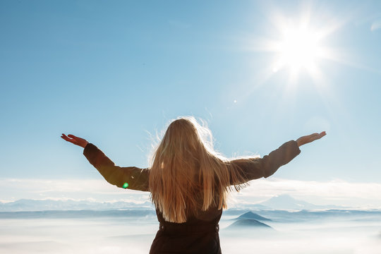 A young blond woman with raised hands enjoys the fresh mountain air and beautiful views of the mountains.