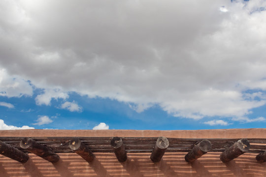 Adobe roofline with exposed vigas and open latillas, blue sky with clouds, copy space, horizontal aspect