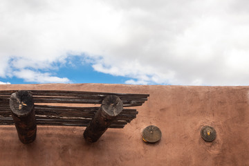 Exterior detail of red adobe building with beams, patch of blue sky with clouds, horizontal aspect